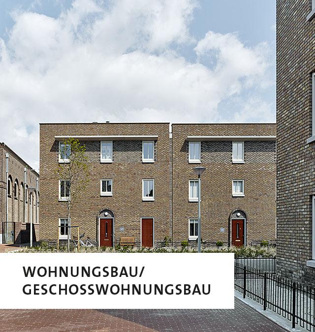 Ein Mehrfamilienhaus mit dunkelbraunem Backstein, links unten die Aufschrift: Wohnungsbau/Geschosswohnungsbau